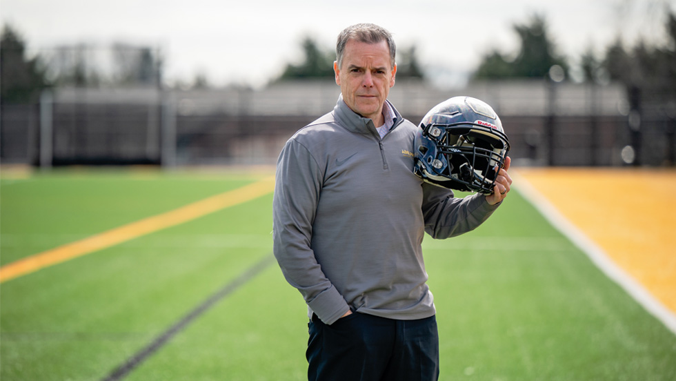 A man standing on a football field holding a football helmet in his left hand. He is wearing a grey quarter-zip pullover and dark pants. The field has green turf with yellow and black markings.