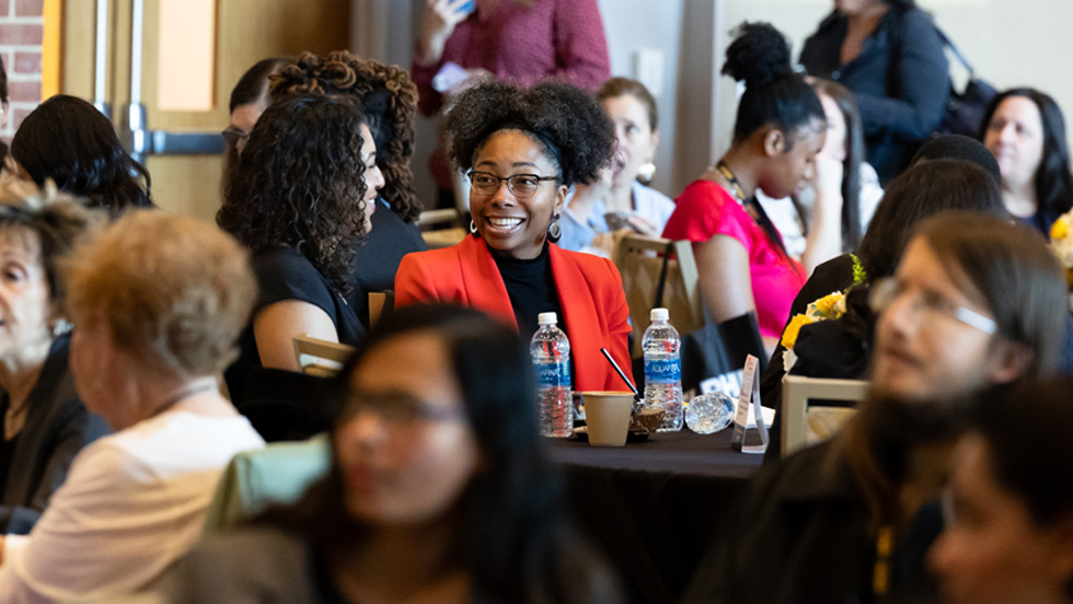 Natalie Ward, wearing a red blazer, sits at a table at the conferences while talking with another student. The two are surrounded other students at tables.