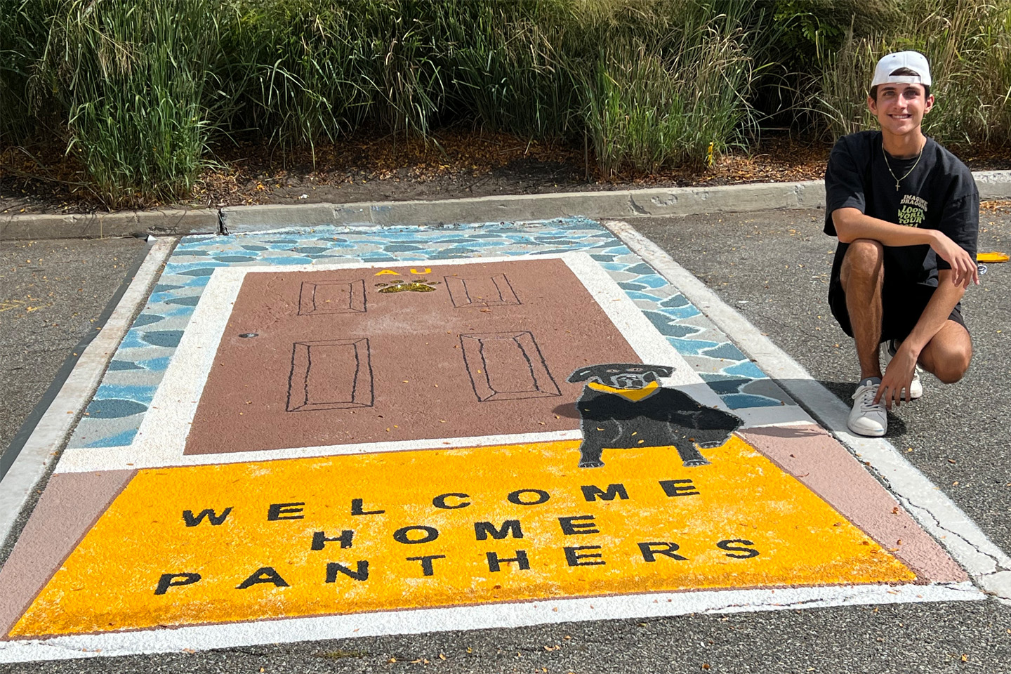 Tullo kneels next to his award-winning design, which shows a welcome mat and black labrador in front of a door labelled "AU." The welcome mat says "Welcome Home, Panthers."