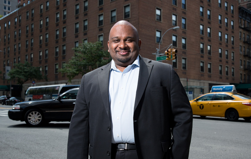 A man wearing a dark suit and light blue dress shirt stands smiling on a city street, with taxis and other cars passing behind him and tall brick buildings in the background.