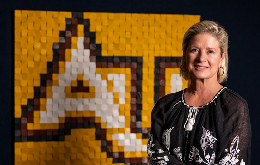 A woman wearing a black blouse with a white floral pattern poses smiling in front of a large, pixelated yellow, black, brown and white artwork featuring the letters “AU."