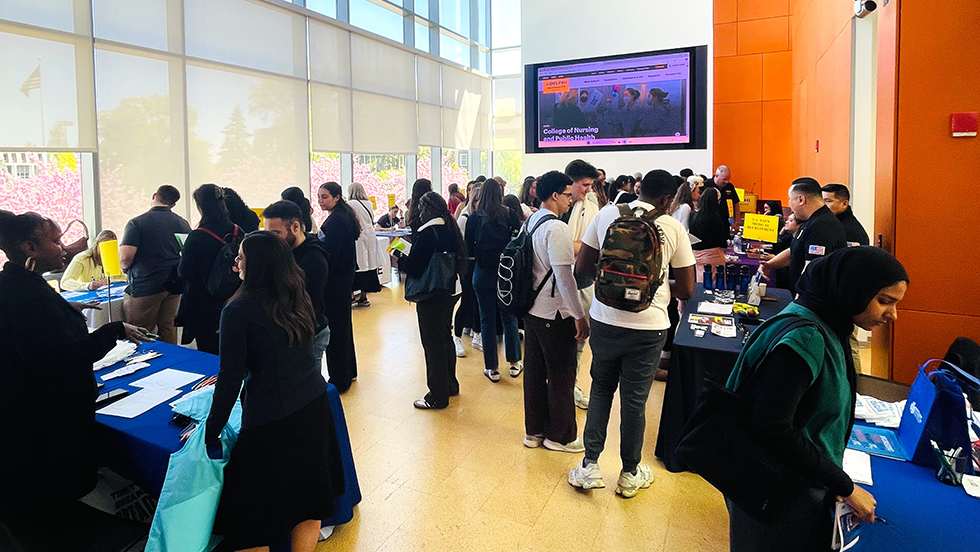 A well-lit, busy event is taking place in a modern building with large windows. A diverse group of young adults and professionals, many dressed in dark business casual attire, are gathered around several tables with blue tablecloths. A large digital screen on the back wall displays a website for a "College of Nursing and Public Health" event. The gathering appears to be a career fair or information session focused on the nursing and public health workforce.