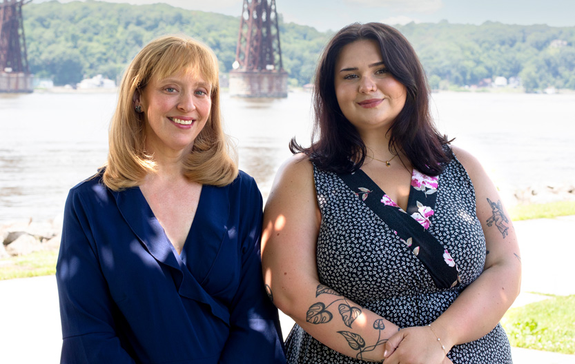 Two women standing side by side outdoors near a waterfront, smiling at the camera. The woman on the left wears a dark blue blouse, and the woman on the right wears a floral sleeveless dress and has tattoos on her arms.