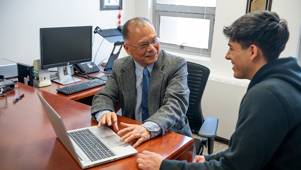 An older man in a gray blazer and blue tie sits at a desk with a laptop, smiling and talking with a younger man in a black hoodie in an office setting