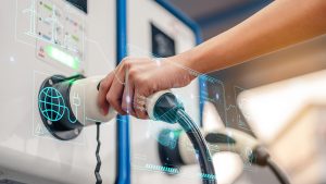 Close-up of a person’s hand plugging a charging cable into an electric vehicle charging station, with digital graphics overlay showing renewable energy and battery system icons.
