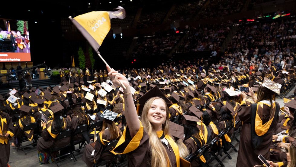 A smiling graduate in a cap and gown stands among a large crowd of fellow graduates in brown and gold seated at a commencement ceremony, waving a flag that reads “Class of 2024.”