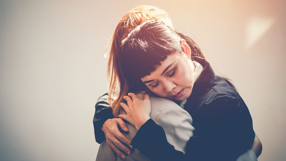 A close-up image of two people hugging each other in a solemn manner. One person has dark hair with bangs and a dark jacket and the other has long hair in a light-colored outfit