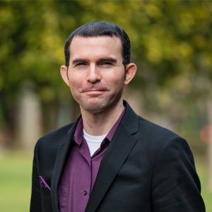 A man with short dark hair smiling wearing a purple shirt and black blazer 