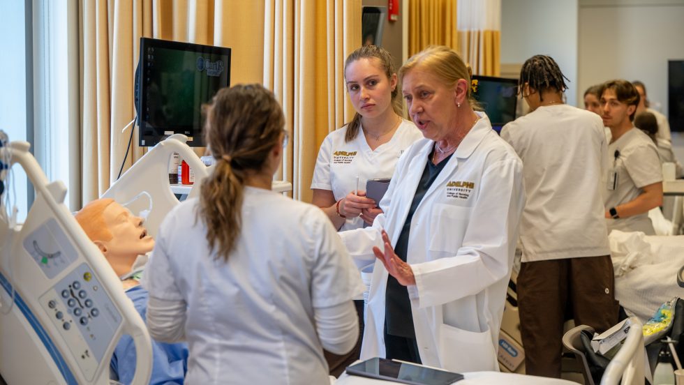 Nursing students in white uniforms training with a simulation manikin.