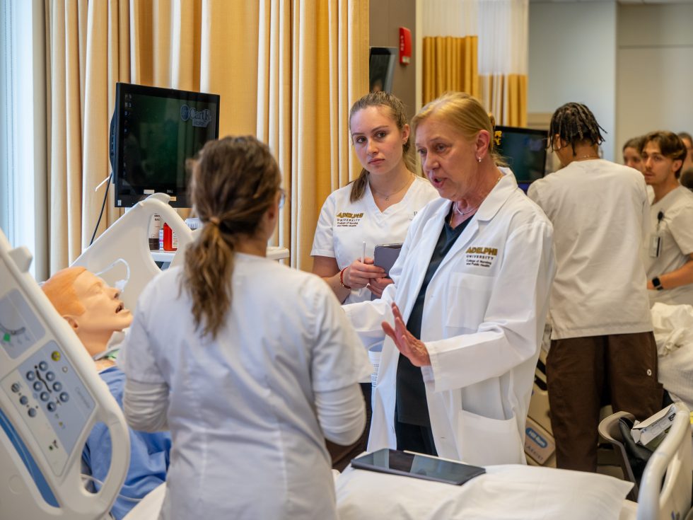 Nursing students in white uniforms training with a simulation manikin.