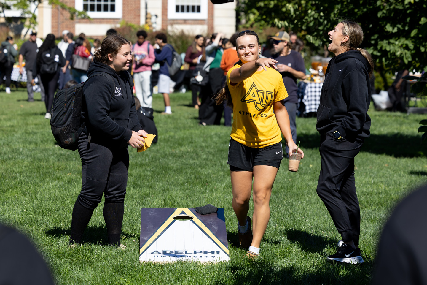 A student plays a game of corn hole. Two other students are at her side, and with a crowd of students in the background.