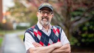 Smiling man with glasses, a beard and a baseball cap standing outdoors with arms crossed, wearing a striped shirt and an argyle sweater vest