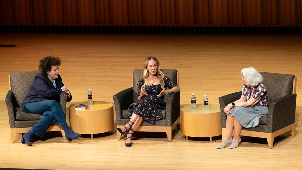 Three women sit on stage engaged in a panel discussion. The woman in the center speaks with her hands raised slightly, while the women on the left and right listen. Two small round tables between them hold bottled water.