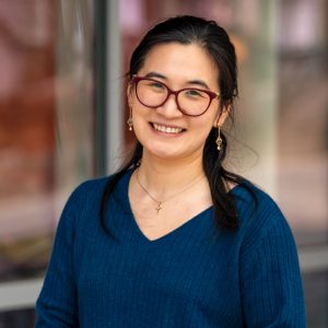 A woman with glasses and long dark hair in a blue sweater smiling