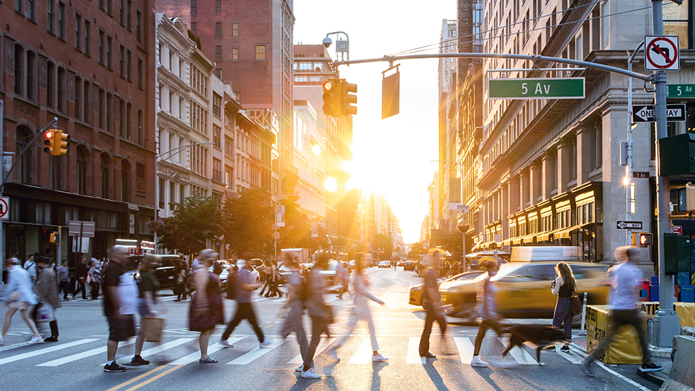 Working professionals cross Fifth Avenue in a busy New York City street scene.