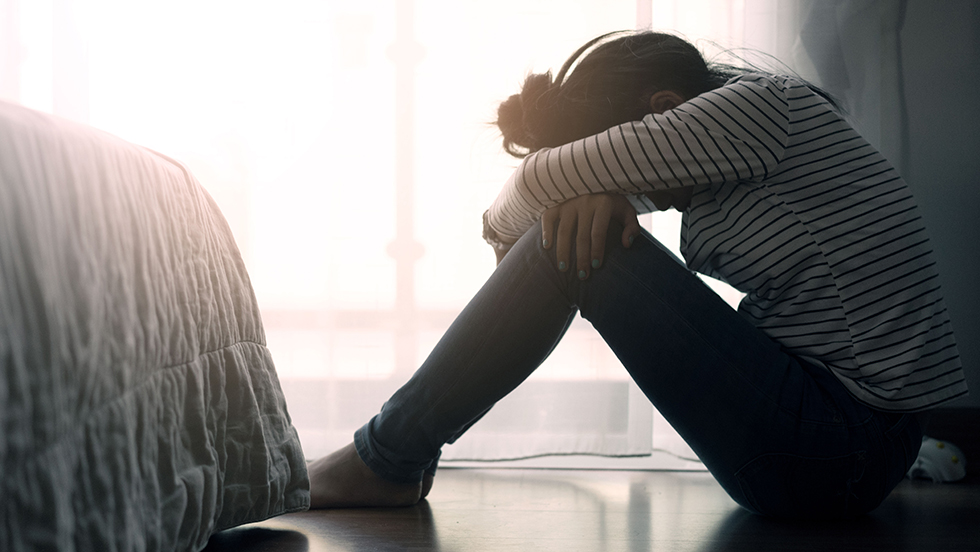 A female sitting on the floor of a bedroom with her knees pulled up to her chest, arms wrapped over her knees and head bowed down on her arms
