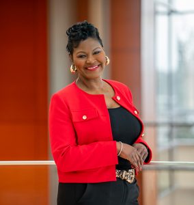 A smiling woman wearing a red blazer over a black outfit and gold hoop earrings with her hands crossed
