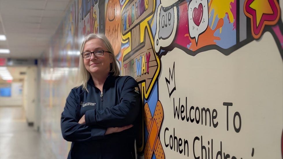The image is a portrait of a woman with light-colored hair and glasses, wearing a dark blue zip-up jacket. She is standing in a hospital-like hallway, leaning against a brightly colored mural that says "Welcome To Cohen Chil..." (presumably Cohen Children's). Her arms are crossed, and she is smiling slightly at the camera. The jacket has a small logo on the left chest area. The background hallway is brightly lit and out of focus.