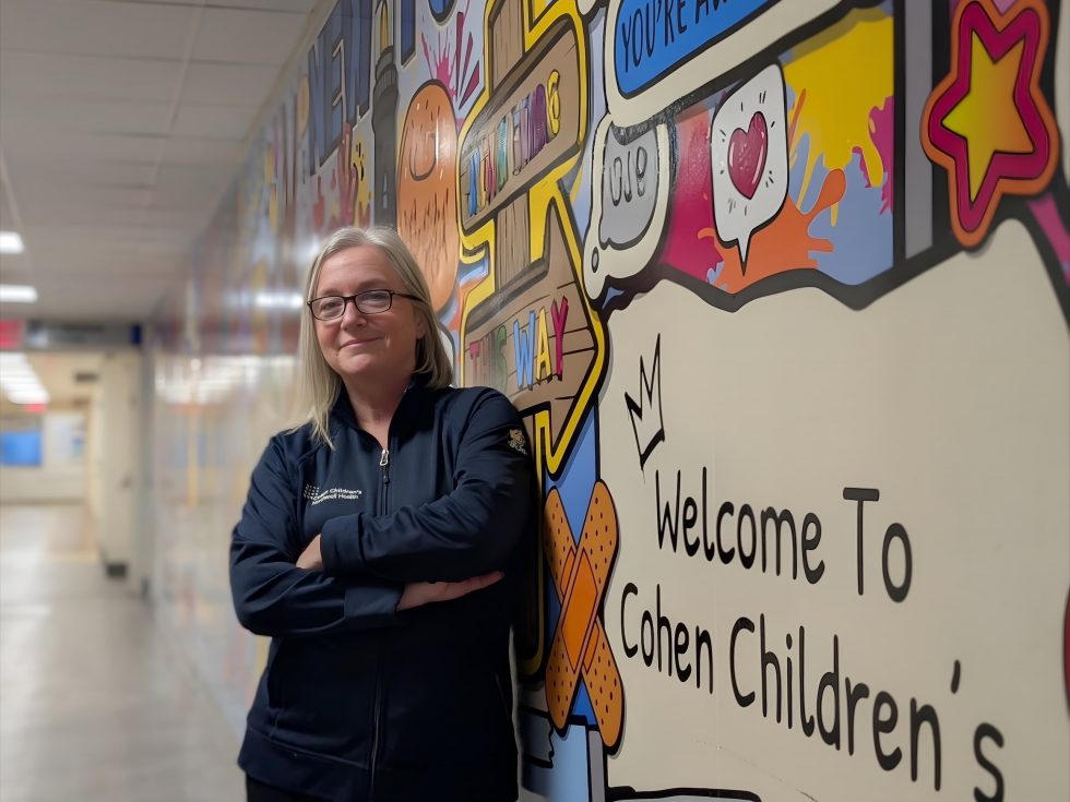 The image is a portrait of a woman with light-colored hair and glasses, wearing a dark blue zip-up jacket. She is standing in a hospital-like hallway, leaning against a brightly colored mural that says "Welcome To Cohen Chil..." (presumably Cohen Children&#039;s). Her arms are crossed, and she is smiling slightly at the camera. The jacket has a small logo on the left chest area. The background hallway is brightly lit and out of focus.