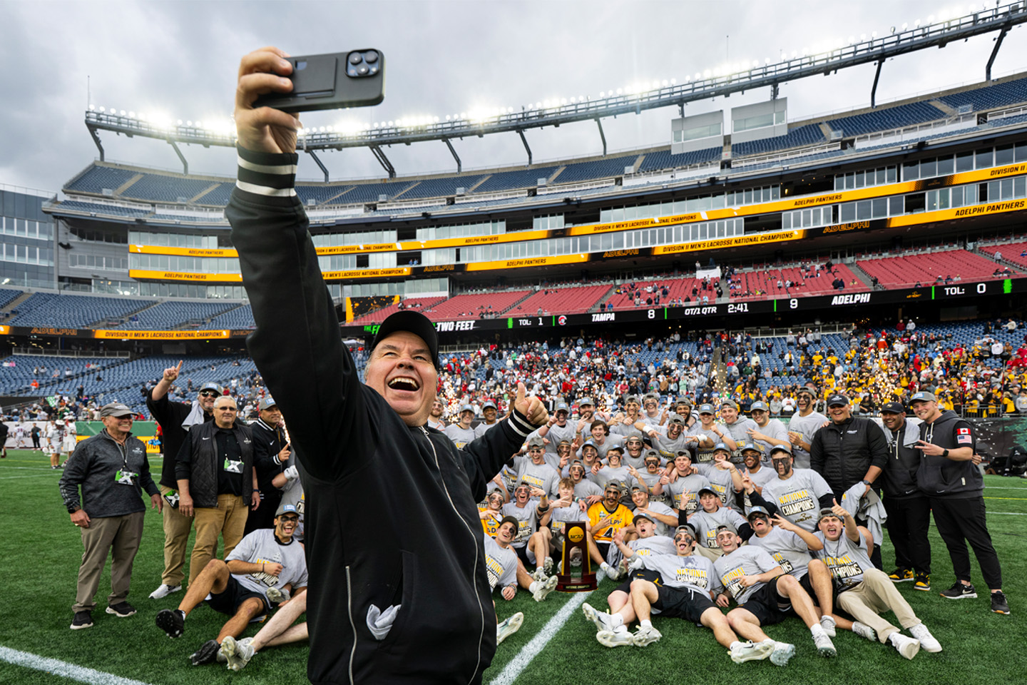 Lacrosse coach Gordon Purdie, in foreground, takes a selfie with the 2025 men's lacrosse team gathered behind him on the ground.
