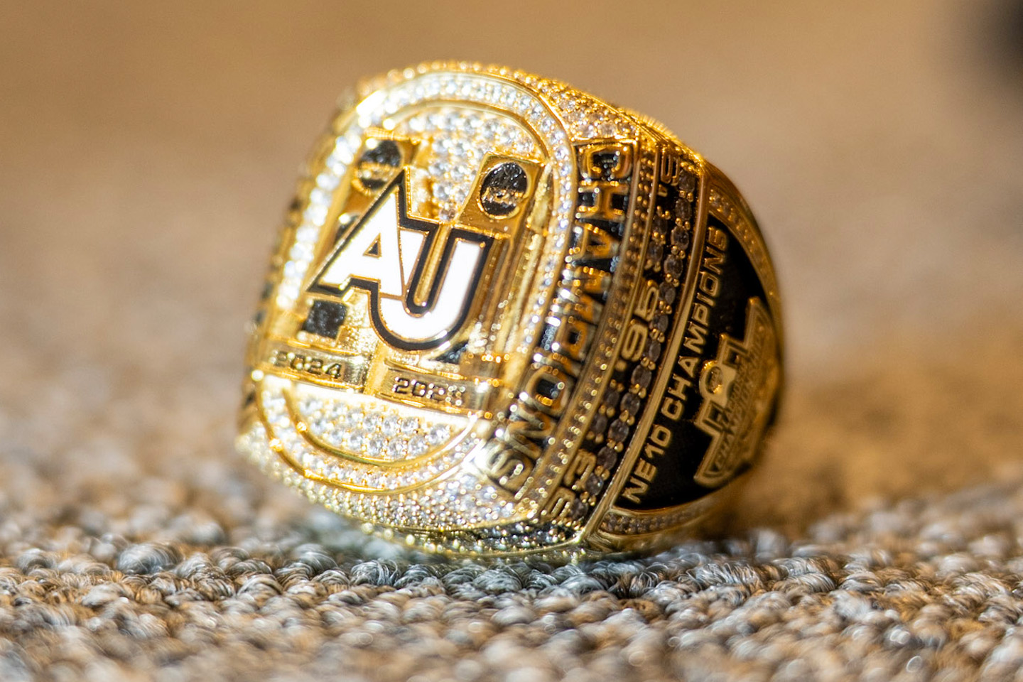 Closeup of a bright gold men's ring, with the letters AU on the front, and the dates 2024 and 2025 inscribed beneath it