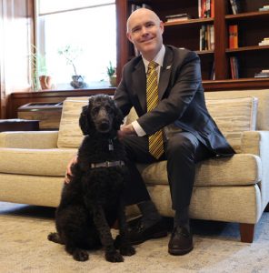 Interim President Christopher Storm, PhD, seated on a couch in his office with a large black poodle at this feet 