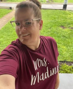 A woman with her hair in a bun and wearing glasses smiles while taking a selfie outdoors. She is wearing a maroon t-shirt that says "Mrs. Madine" in white script.