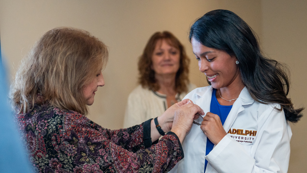 A female professor adds a pin to a female student’s white coat as the associate director of field and clinical experiences looks on. They look proud and happy.