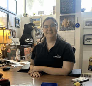 A smiling woman in an Adelphi shirt sits at a cluttered desk filled with colorful and encouraging messages, notes and pictures