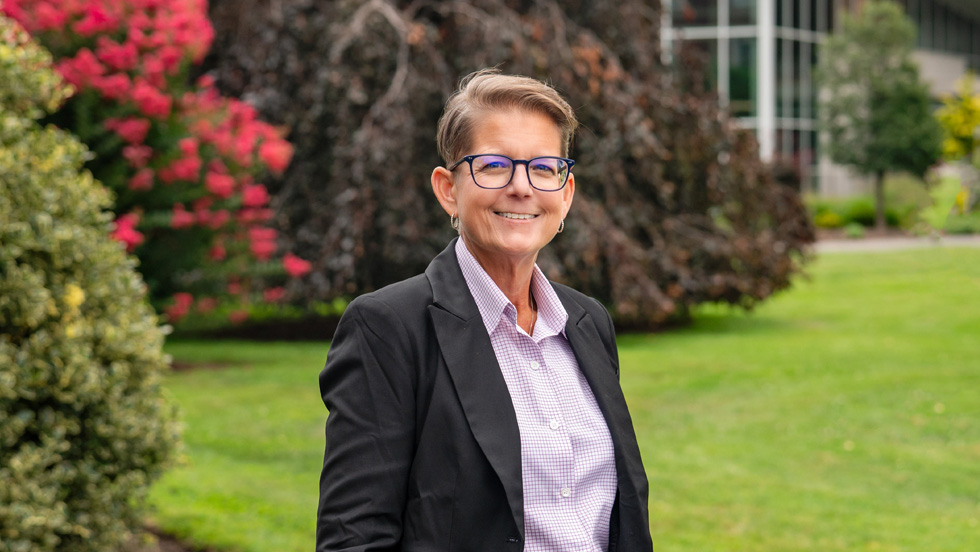 A smiling, middle-aged woman with short, light brown hair and glasses, wearing a black blazer over a light purple patterned button-down shirt. She is standing outdoors on a grassy lawn with dark, reddish-purple leafed trees and bright pink/red flowering bushes in the background.