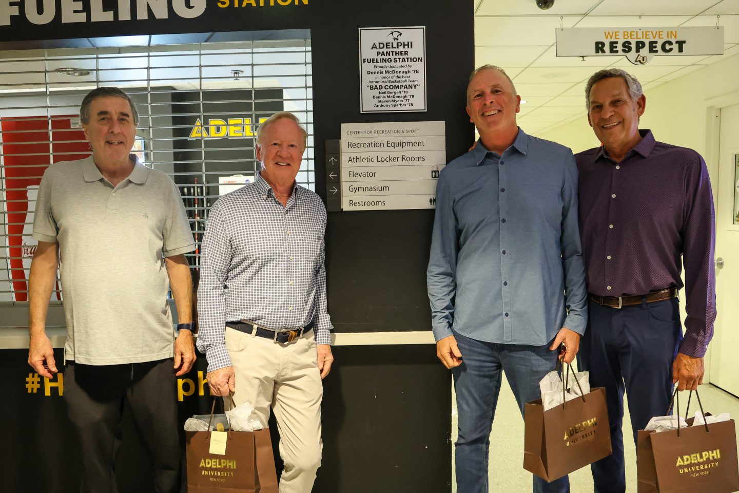 Four adult men stand smiling in front of a wall labeled “Panther Fueling Station” inside a campus recreation center.