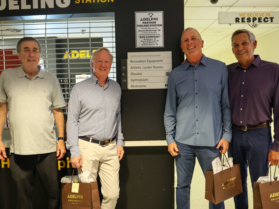 Four adult men stand smiling in front of a wall labeled “Panther Fueling Station” inside a campus recreation center.