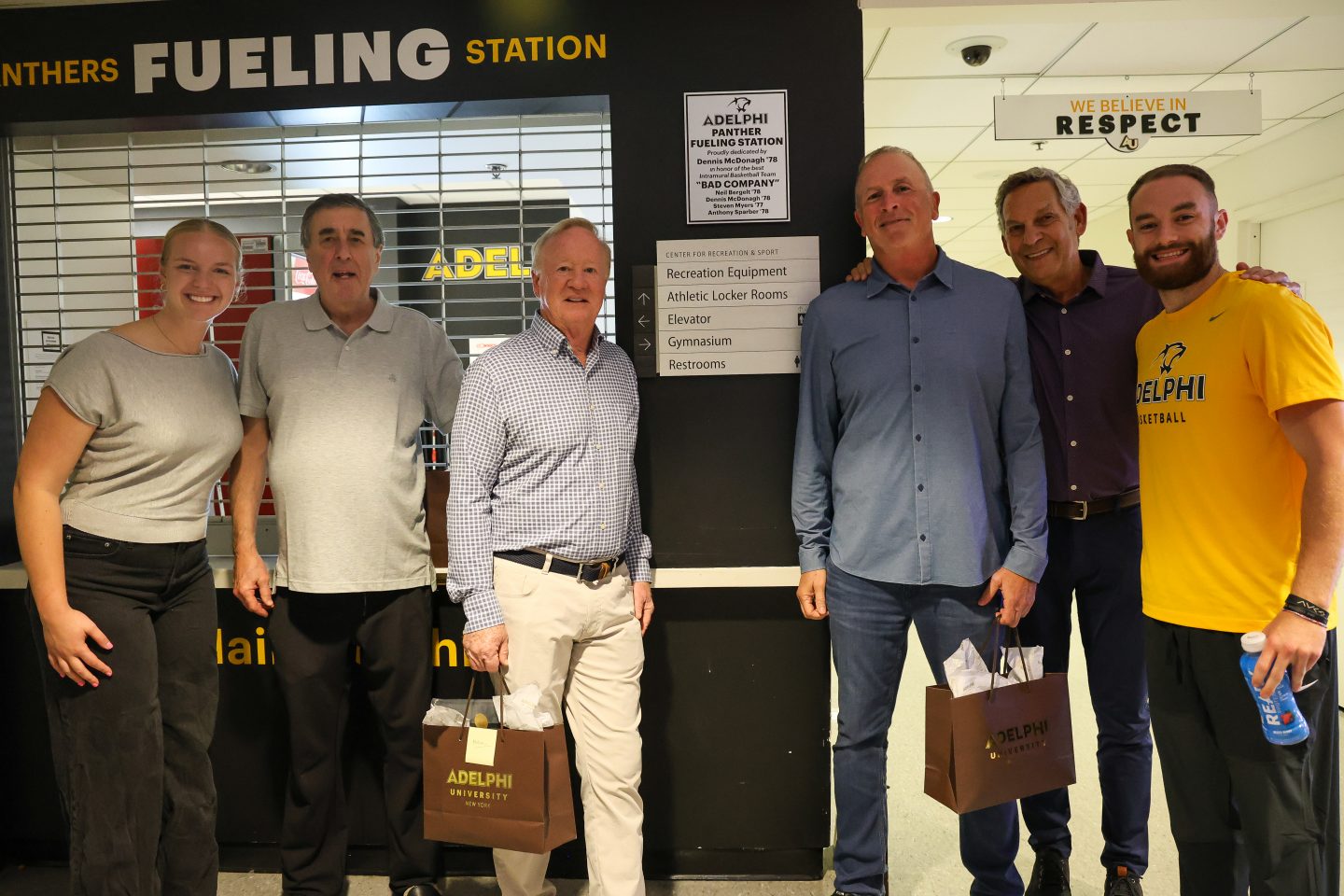Six people stand smiling in front of the Panther Fueling Station, including four adult men and two younger adults wearing athletic apparel.