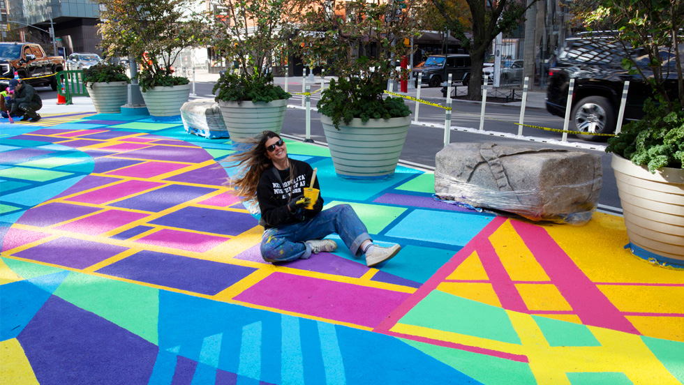 Mural artist Cara Lynch ’12 sits on a colorful painted street, paintbrush and paint can in hand, her hair blowing in the wind, smiling broadly. 