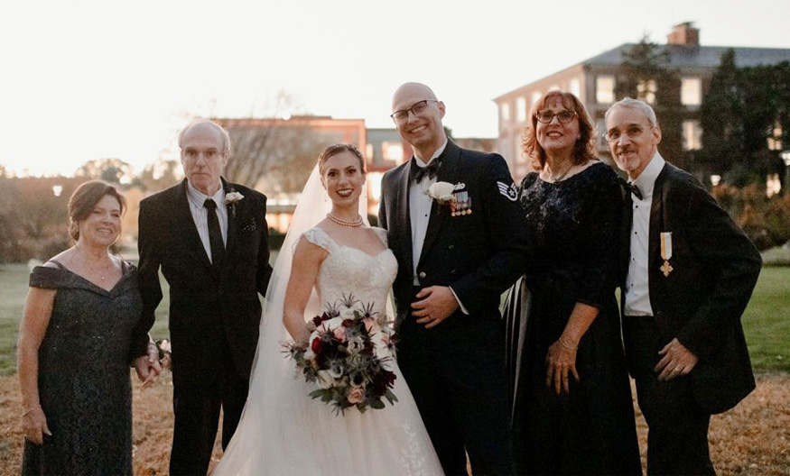 a bride in bridal gown and groom in a tuxedo standing outside on the Adelphi campus at sunset with their mothers and fathers