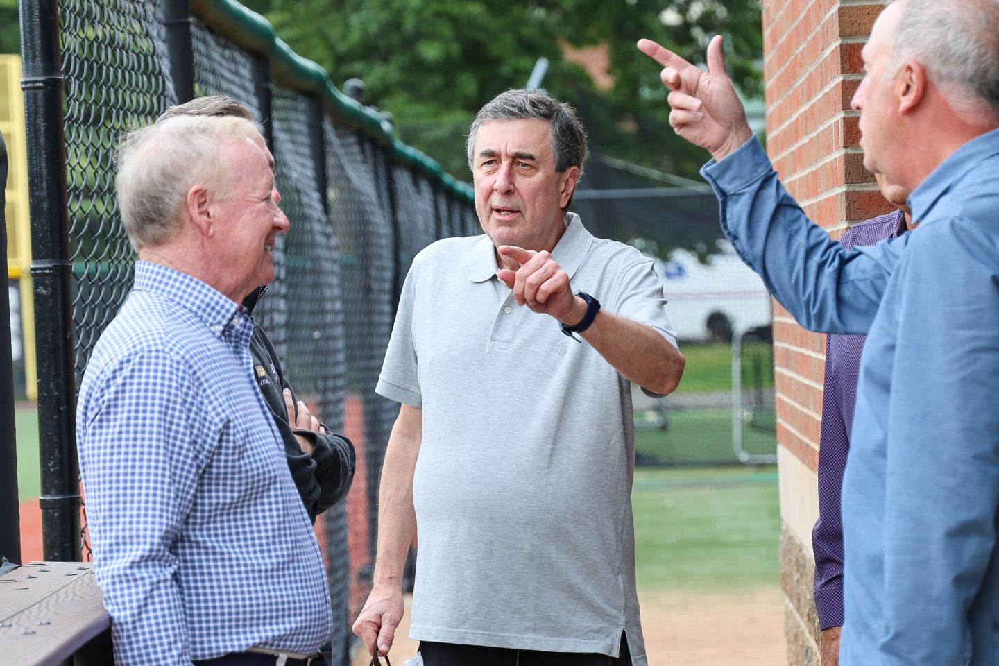 Three adult men stand near a brick wall and chain-link fence, gesturing and talking during an outdoor campus visit.