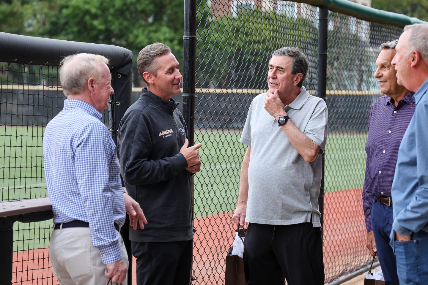 Several adult men stand in a small group beside a chain-link fence, talking and smiling near an outdoor athletic field.