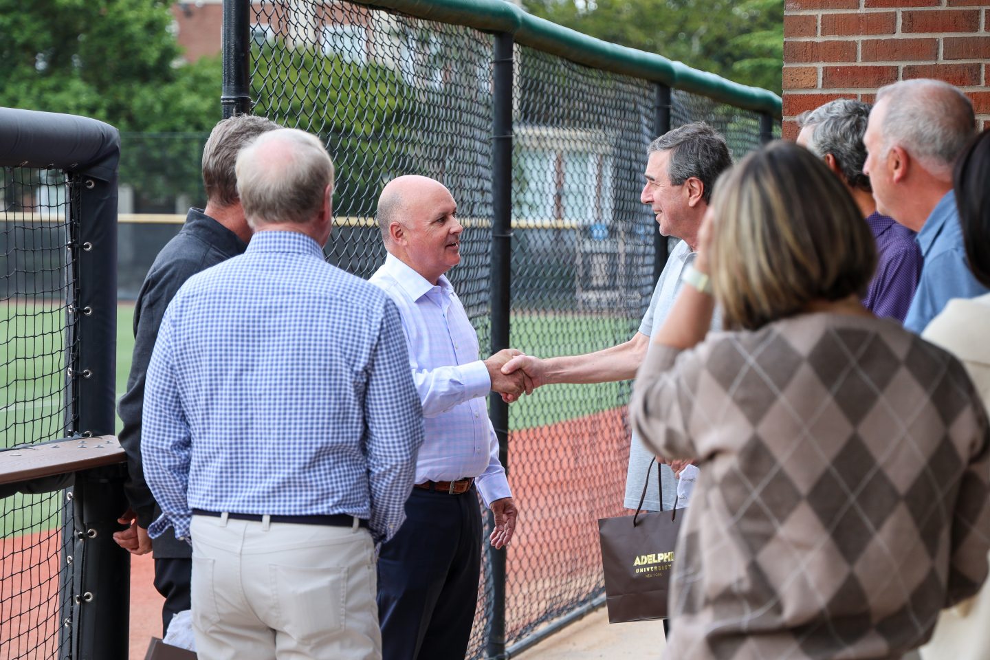 Two adult men shake hands while standing beside a chain-link fence near an outdoor athletic field.