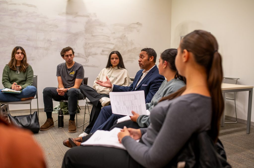 Errol Rodriguez among students sitting in chairs in a rounded formation, male student all the way to the left, then a female student, an older man