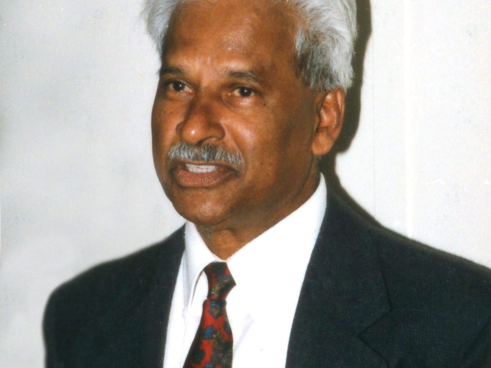 A formal portrait of an older man with silver hair and a mustache, dressed in a charcoal suit, white shirt, and a red and blue patterned tie, looking slightly off-camera.