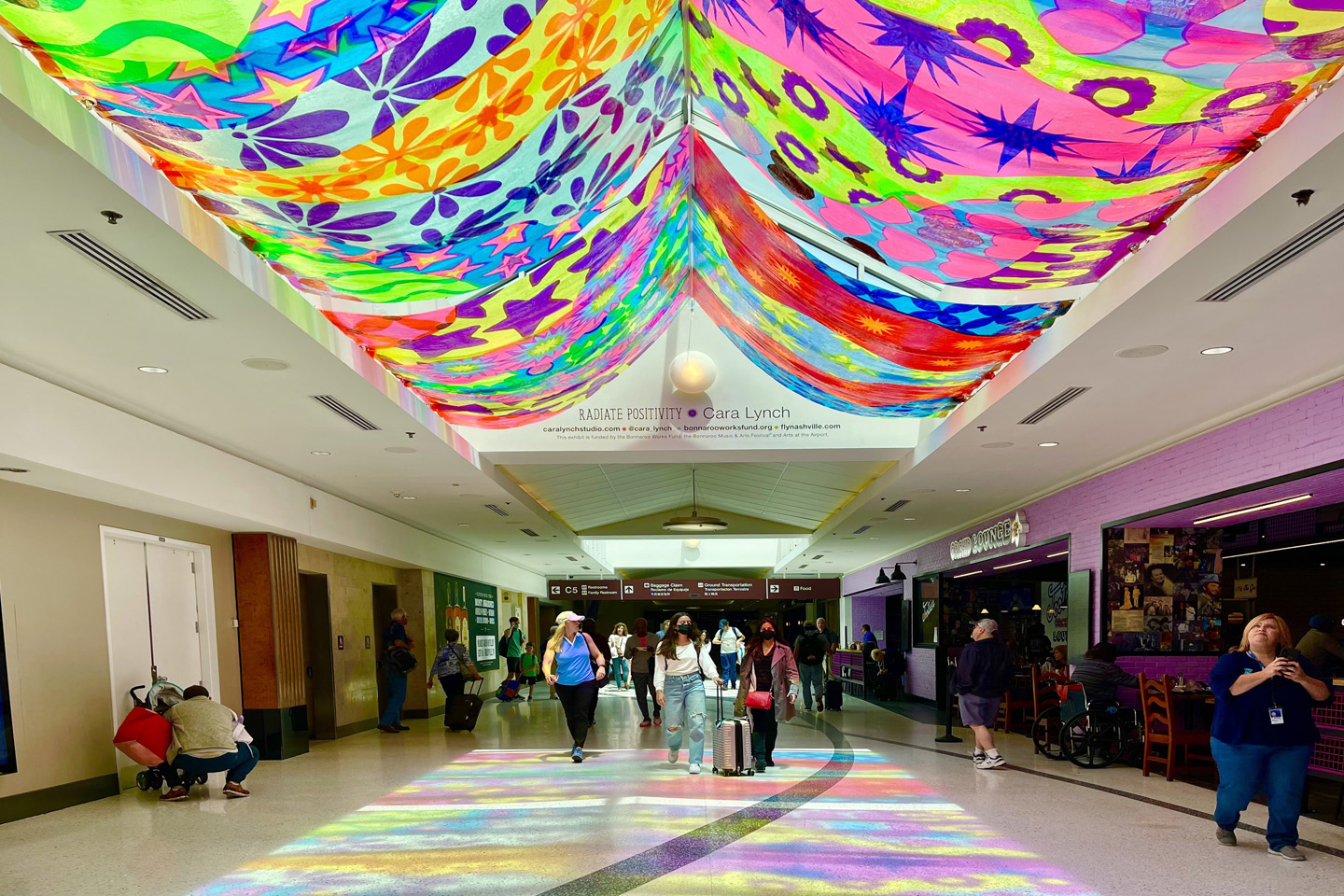 Passengers walk through the airport terminal. The vinyl drapery of Radiate Positivity shines bright colors throughout the passage way and on to the terminal floor.