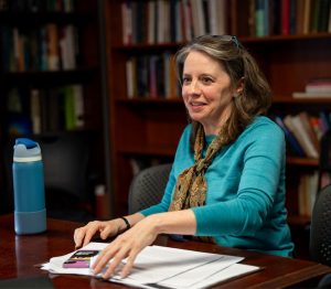 A female professor, with papers on her desk, looks like she is conversing with a student in her office. 