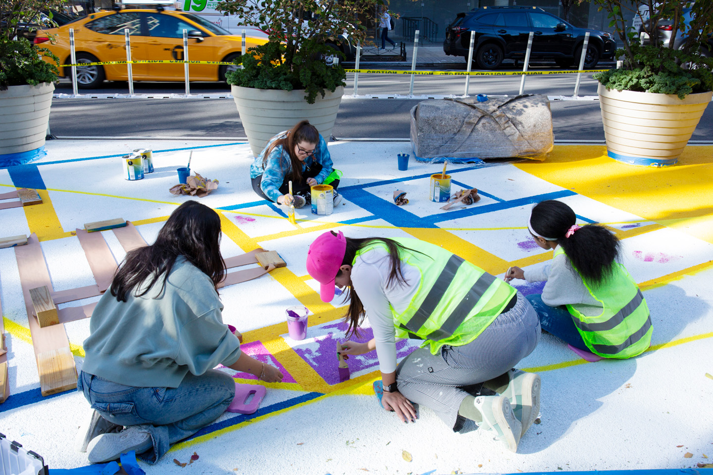 Four students in jeans and tee shirts, paintbrushes in hand, lean over a city street, painting in colorful designs.