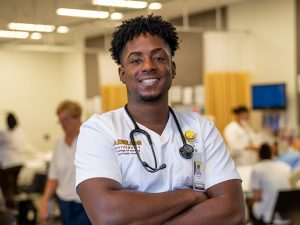 A male nursing student stands proudly in the simulation lab, arms crossed, at the College of Nursing and Public Health.