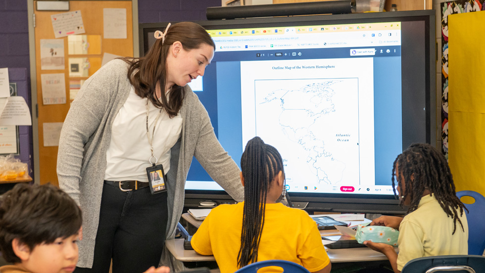 A female teacher leans over a desk to assist two students in a classroom while a map of the Western Hemisphere is displayed on a large digital touchscreen monitor behind them