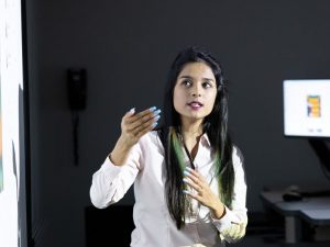 A young woman, professionally dressed, stands in front of a screen, presenting to a class.