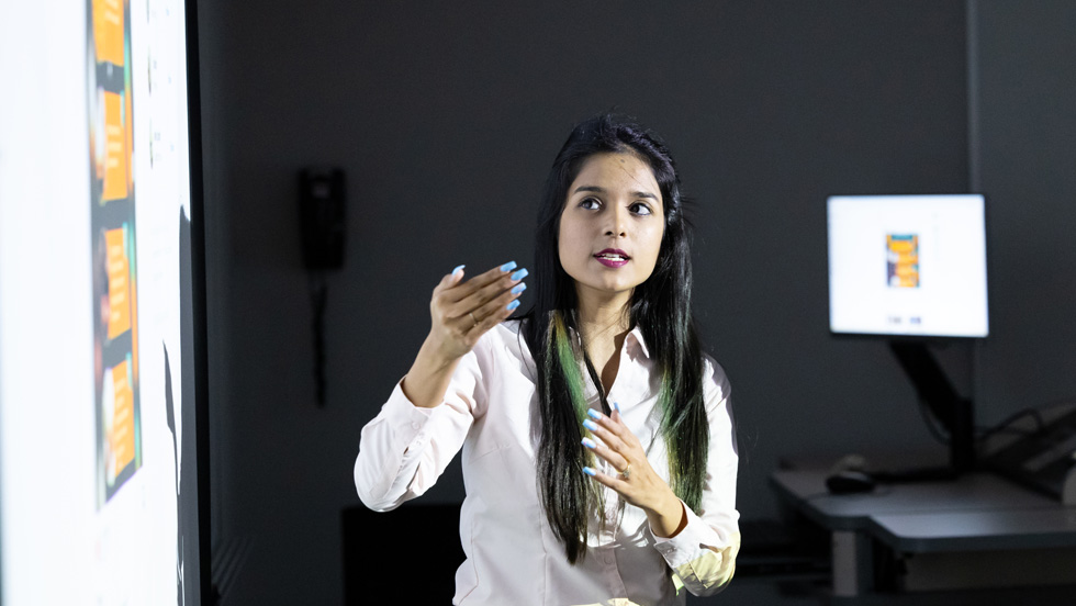 A young woman, professionally dressed, stands in front of a screen, presenting to a class.