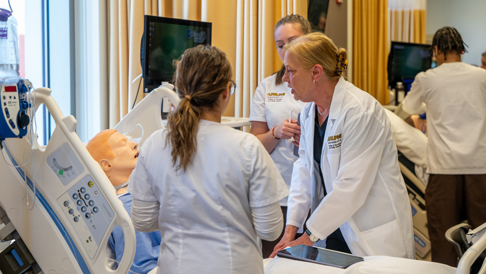 A faculty member and two students at the side of a hospital bed attending to one of the lifelike manikins in the simulation lab.