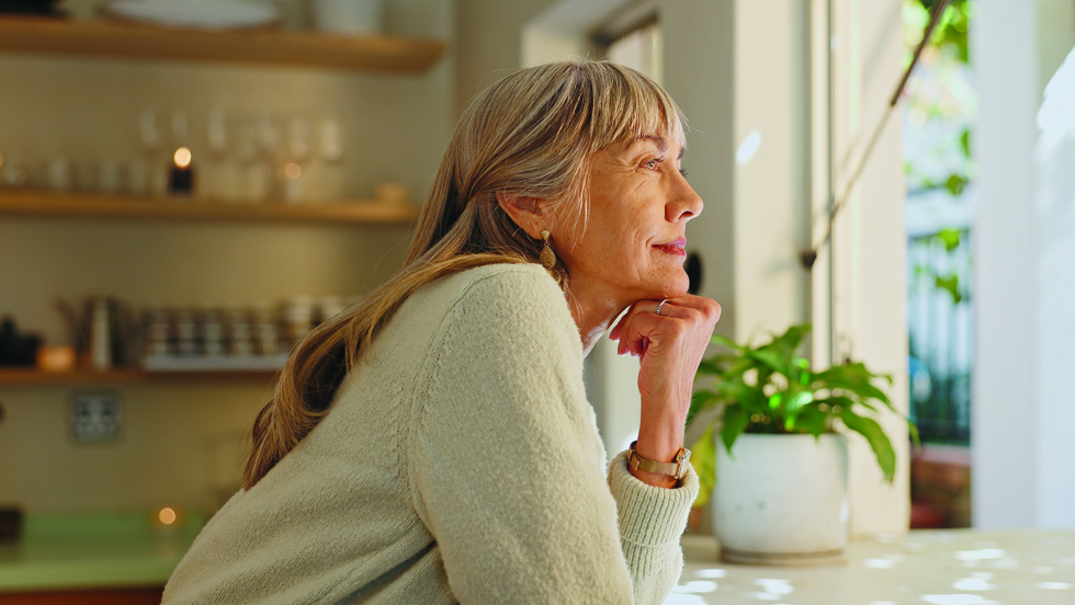 A mature woman with gray hair resting her chin on her hand and looking thoughtfully out a sunlit window