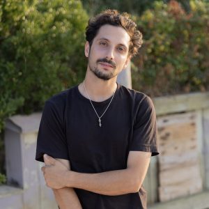 Headshot of Carl Timpone standing outdoors with arms crossed, wearing a black t-shirt and a silver pendant necklace, with greenery and a wooden structure in the background.